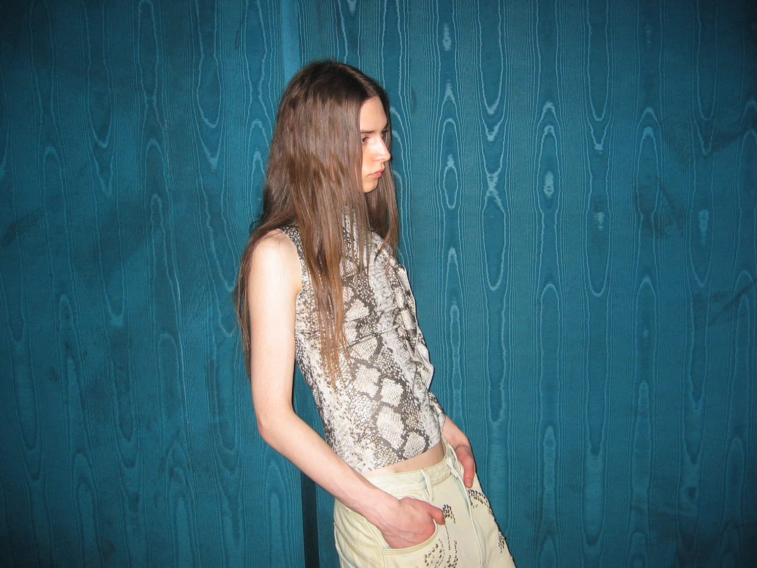 AURA LAND snake-print sleeveless top and pale trousers in profile against a blue wall